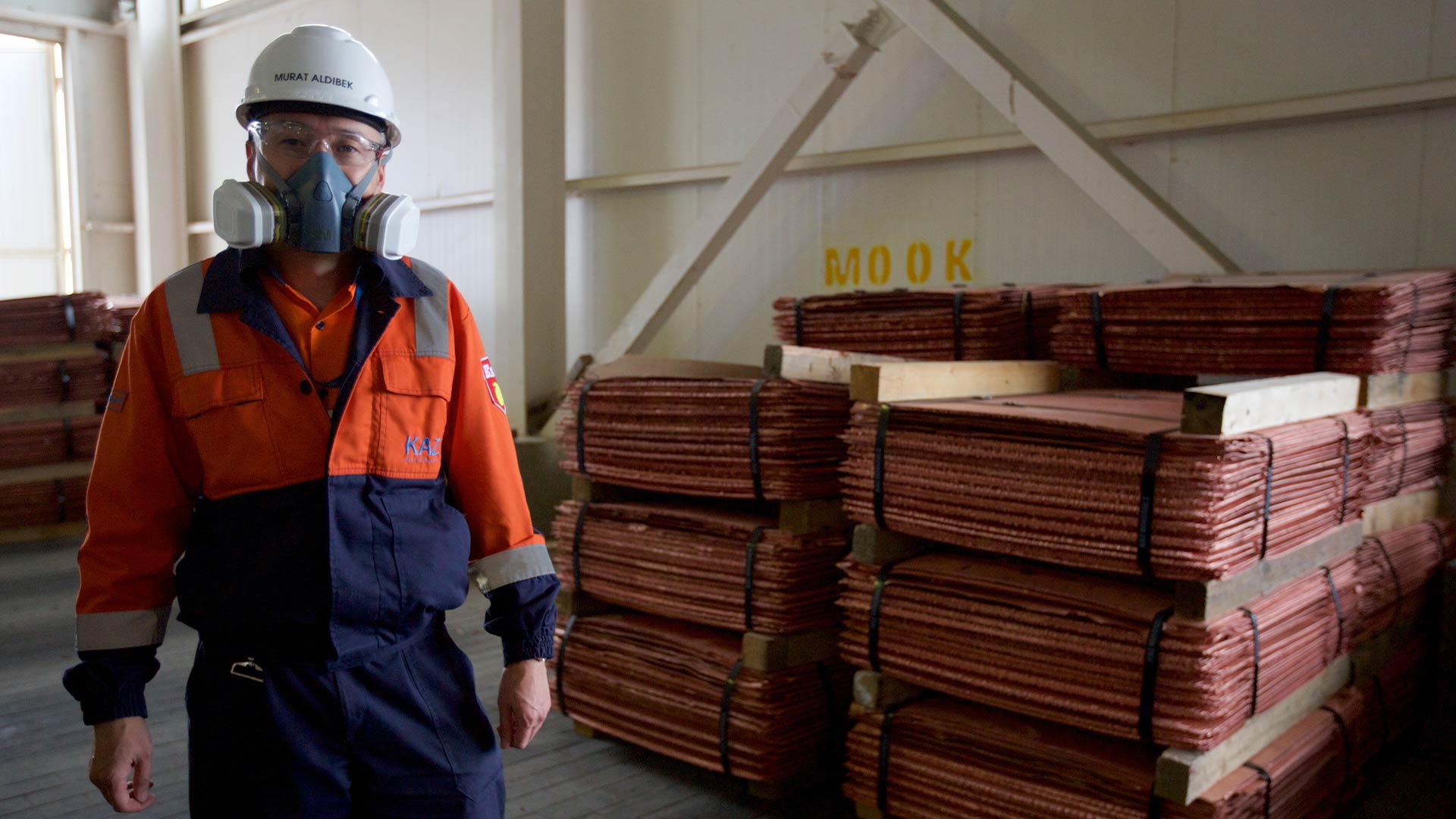 Человек в защитной экипировке смотрит в камеру. На заднем плане сложены материалы.