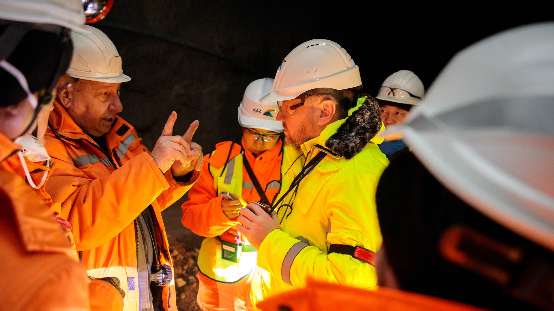 На переднем плане стоят трое рабочих в защитной экипировке и ведут разговор. На заднем плане виден склад и перерабатывающий цех.